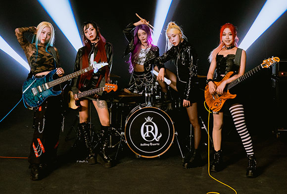 Five women in dark, edgy outfits pose with guitars and drums under bright stage spotlights.