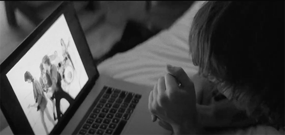 A person's hands clasped near their face while looking at a laptop screen in a dimly lit room.