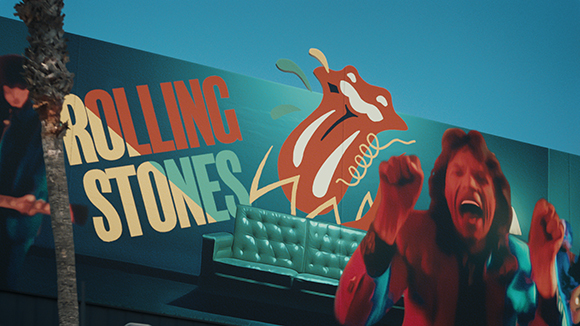 A person with raised fists and an open mouth shouts in front of a large Rolling Stones mural.
