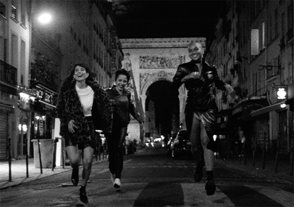 Three people running and laughing down a dark city street toward the camera at night.
