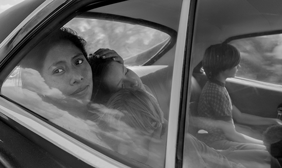Black and white shot looking through a car window at a woman embracing a child and another person nearby.