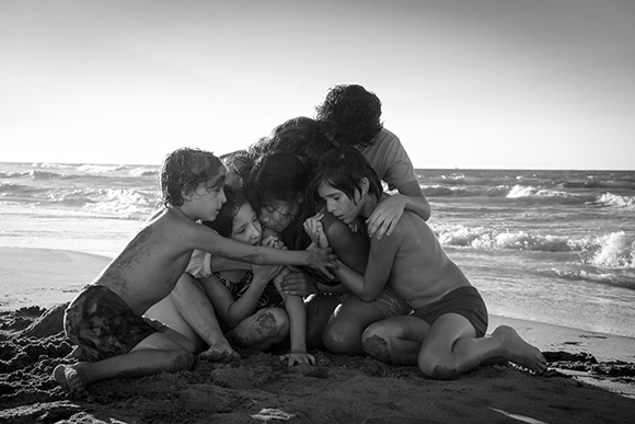 Black and white photo of several children huddled closely together on a sandy beach by the ocean waves.