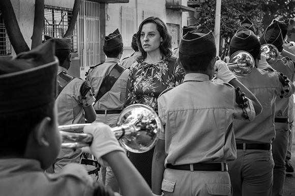 A woman in a floral blouse stands surrounded by young musicians in uniform playing brass instruments.