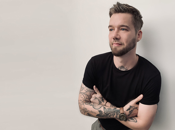 A bearded man with arm and neck tattoos wearing a black t-shirt, looking away from the camera.