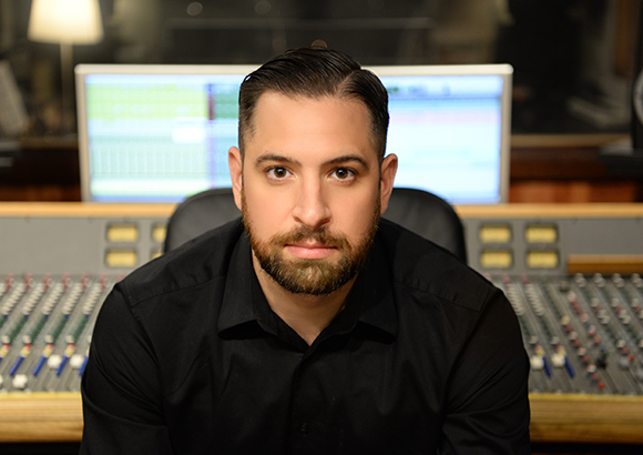 A man with dark hair and a beard wearing a black shirt sits before a large audio mixing console.