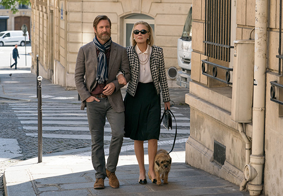 A man in a blazer and a woman in a patterned jacket walk a small dog down a sunny city street.