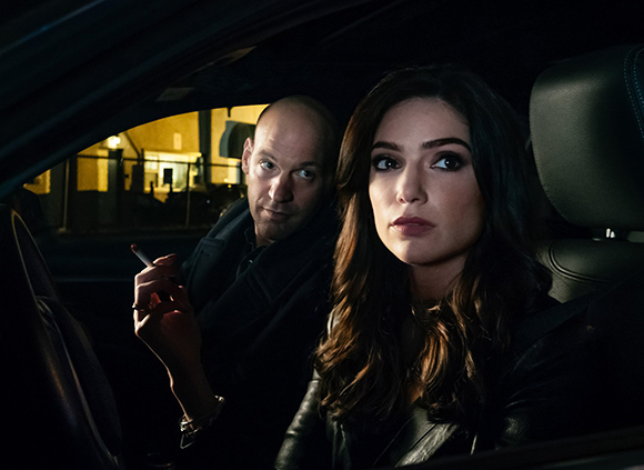 A woman with long dark hair looks ahead while a bald man sits behind her holding a cigarette.