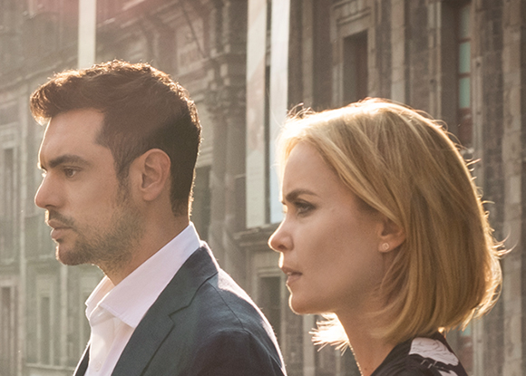 Profile view of a man in a suit and a blonde woman looking toward the left against a city background.