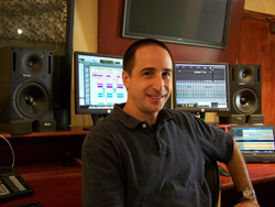 A man smiling while sitting at a desk with multiple computer monitors and studio speakers.