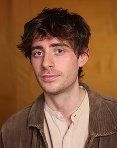 A man with dark, messy hair and blue eyes looking toward the camera against a yellow background.