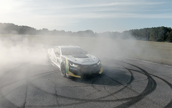 A white and yellow race car drifts around a curve on an asphalt track, creating thick clouds of white smoke.