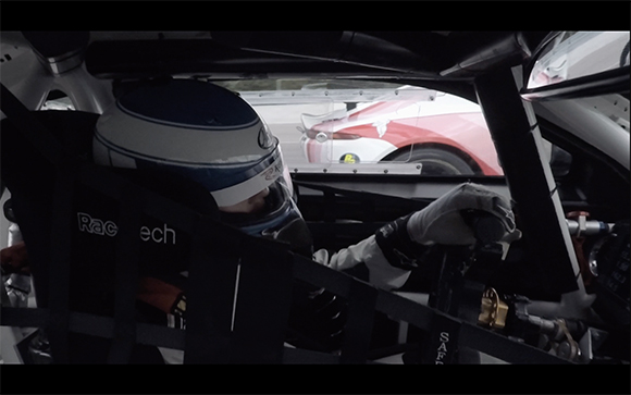 A driver wearing a blue and white helmet and white gloves sits inside a race car cockpit.
