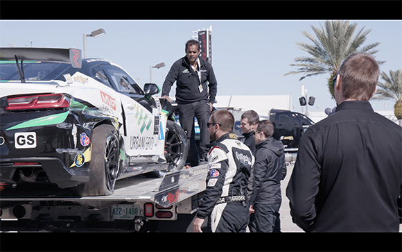 A white and black race car sits on a flatbed trailer as crew members stand around it.