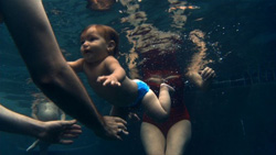 A baby in a blue diaper swims underwater near an adult in a red swimsuit.