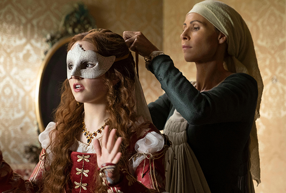 A woman with long red hair wears a white masquerade mask while another woman adjusts her veil.
