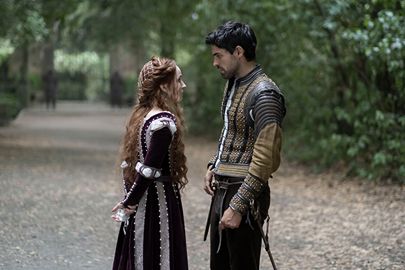 A man in gold and black armor stands facing a woman in a long purple velvet dress on a forest path.