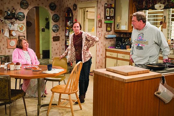 A woman in a pink robe sits at a table while another woman stands nearby and a man stands in the kitchen.