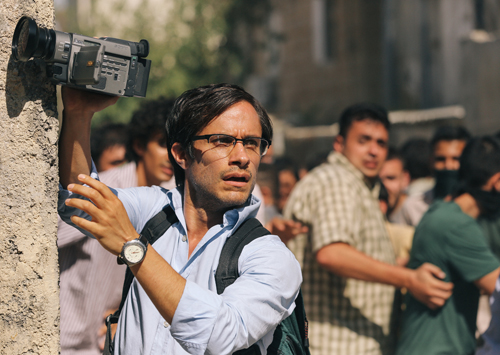 A man in a light blue shirt holds a video camera up to film a crowd in an outdoor urban area.