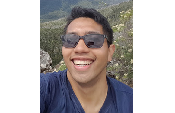 A smiling man wearing dark sunglasses and a navy blue t-shirt in a natural setting.