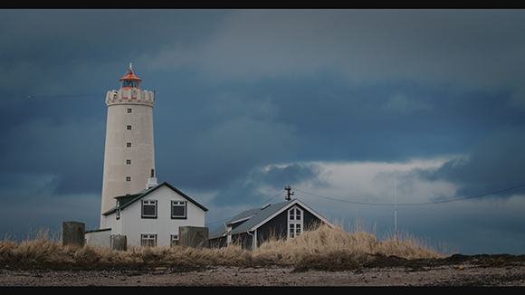 A tall white lighthouse tower rises behind small dark houses under a heavy, overcast sky.