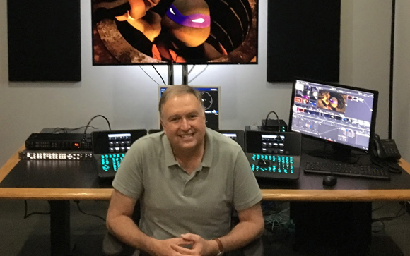 A man sits at a desk with multiple computer monitors and editing equipment in a studio.