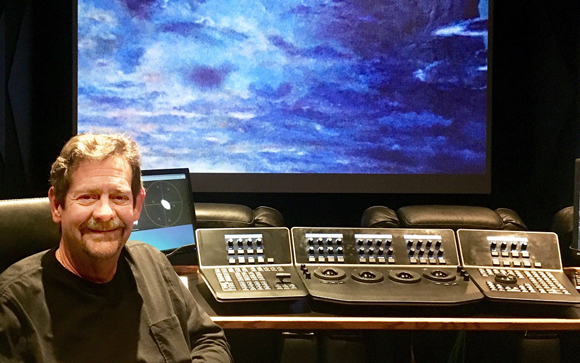 A man sits in a dark studio in front of a large blue screen and a professional color grading control panel.