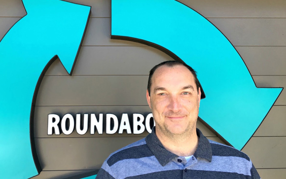 A man in a striped polo shirt stands in front of a large teal circular arrow logo and the word Roundabout.