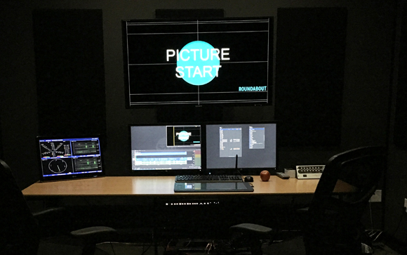A desk with three computer monitors displaying video editing software and a 'PICTURE START' graphic.