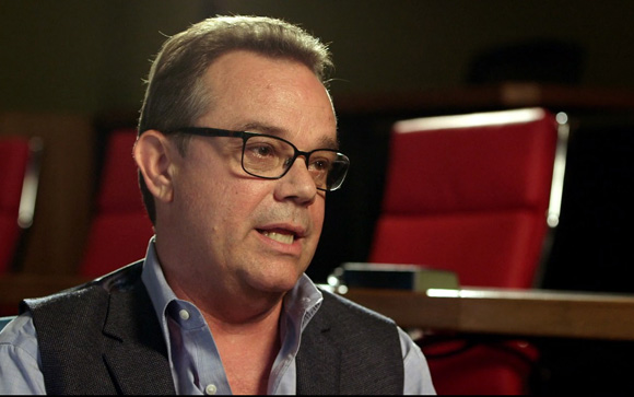 A man with glasses and a dark vest speaks in a room with red chairs.