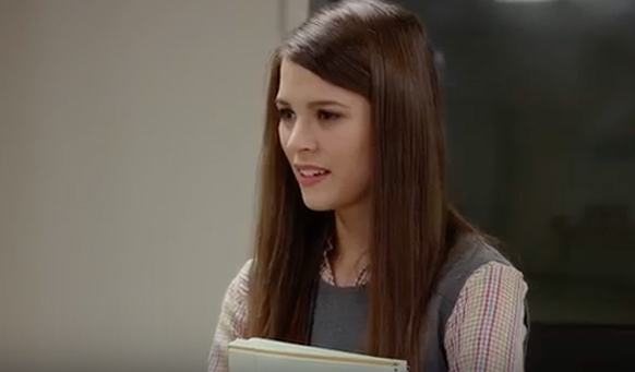 A young woman with long brown hair wearing a grey vest over a patterned shirt.