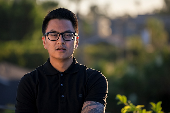 A man with dark hair and glasses wearing a black polo shirt outdoors during golden hour.