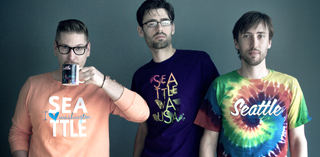 Three men stand against a gray wall wearing shirts with Seattle text and tie-dye patterns.