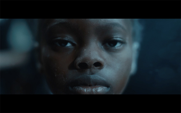 Close-up of a person's face looking directly at the camera in low, blue-toned light.