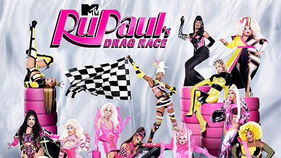 A group of drag performers in vibrant, stylized costumes pose against a white draped background.