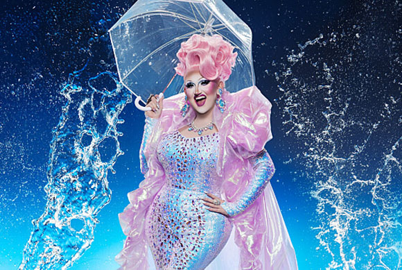 A person in a sparkling pink gown and large pink wig holds a clear umbrella amidst splashing water.