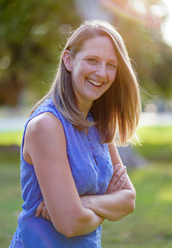 A smiling woman with light brown hair wearing a blue sleeveless top, posing with arms crossed outdoors.