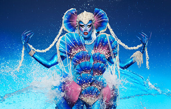 A person in a blue and pink seashell costume with pearl strands against a blue background with water splashes.