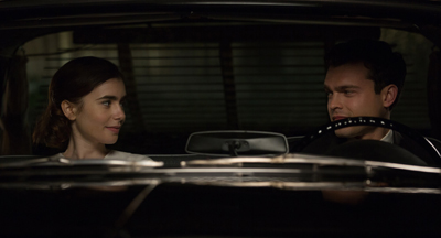 A man and a woman sit in a dark car, looking at each other.