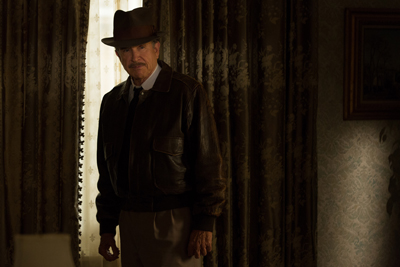 A man wearing a brown fedora, leather jacket, and tie stands in a dimly lit room near a window.