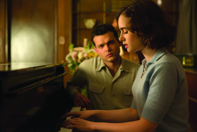 A woman in a light blue polo shirt plays a piano as a man in a tan shirt watches her from behind.