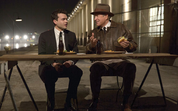 A man in a suit and a man in a leather jacket and hat sit at a wooden table eating outdoors at night.