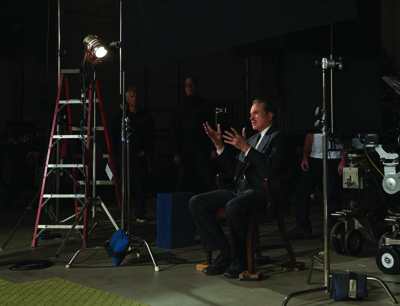 A man in a dark suit sits in a wooden chair on a dimly lit film set, gesturing with his hands.