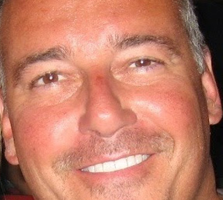 Close-up of a man with short grey hair and a mustache smiling at the camera.