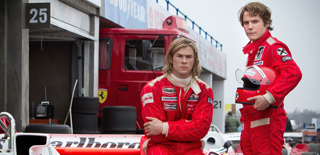 Two men in red racing suits stand in front of a red truck and a racing garage.