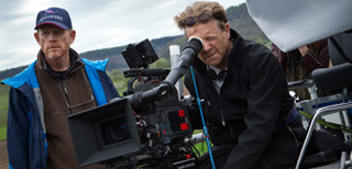 Two men operate a large professional film camera on a tripod in an outdoor setting.