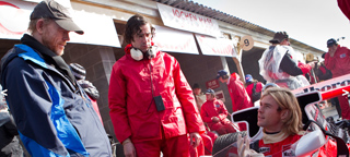 Pit crew members in red and blue jackets standing near a racing vehicle in a garage.