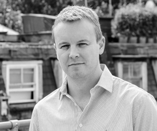 A man with short hair wearing a light-colored button-down shirt, looking forward against a blurred outdoor background.