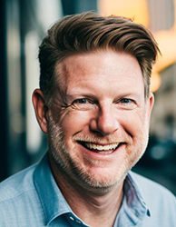A smiling man with reddish-brown hair and a light beard wearing a light blue button-down shirt.