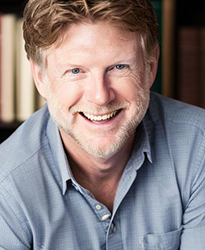 A smiling man with light hair and a beard wearing a blue button-down shirt.