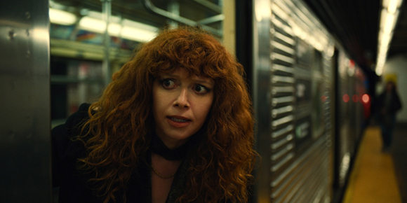 A woman with voluminous curly red hair looks toward the camera with a wide-eyed, startled expression in a subway.